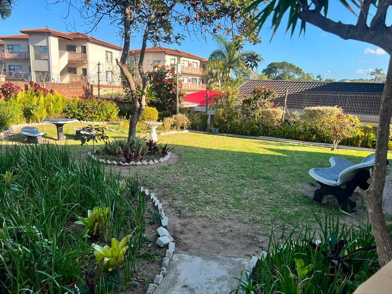 A well-maintained communal garden area featuring established trees, landscaped flower beds, and a concrete pathway within a secure apartment complex setting.