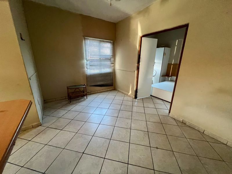 An empty living area featuring tiled flooring and a window with venetian blinds, leading into an adjacent bedroom space.