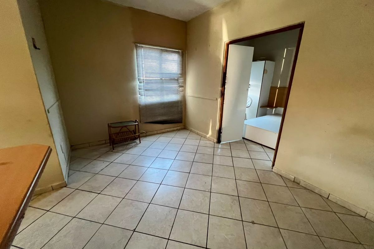 An empty living area featuring tiled flooring and a window with venetian blinds, leading into an adjacent bedroom space.