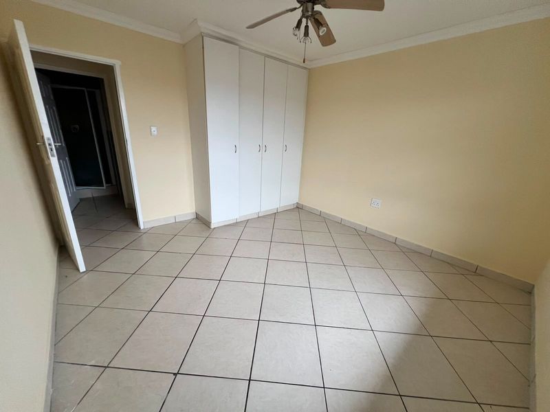 A clean, empty bedroom featuring tiled flooring, built-in cupboards, and a ceiling fan, with an adjacent doorway leading to another room.