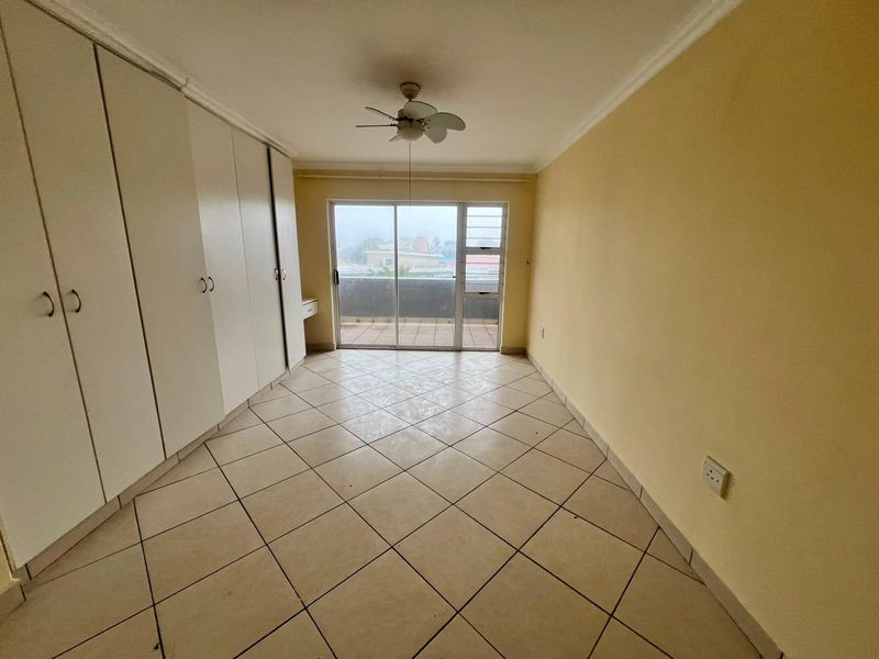 A bright, empty bedroom featuring tiled flooring, extensive built-in cupboards, a ceiling fan, and direct access to a private balcony through sliding glass doors.