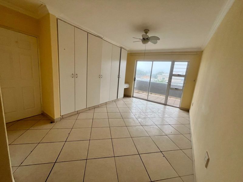 A spacious, empty bedroom featuring built-in cupboards, tiled flooring, a ceiling fan, and direct access to a private balcony through sliding glass doors.