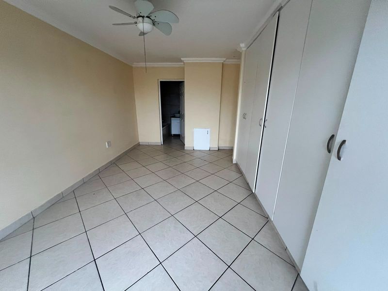 A spacious, empty bedroom featuring neutral tiled flooring, built-in cupboards, and a ceiling fan, with direct access to an adjoining bathroom.