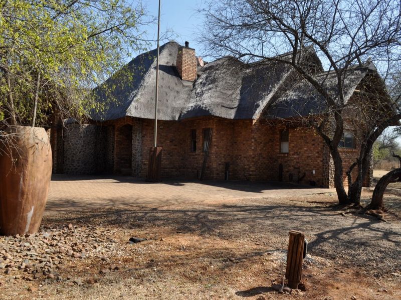 A rustic house featuring a traditional thatch roof, brick and stone facade, and a paved driveway set in a natural landscape.