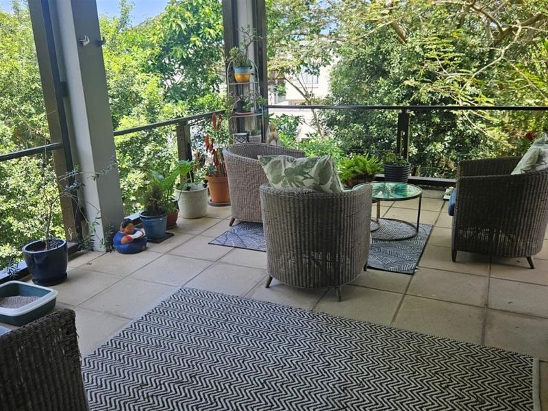 A covered balcony featuring tiled flooring and a glass balustrade, offering an elevated outdoor space with views of surrounding trees.