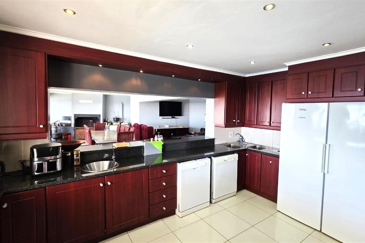 A functional kitchen featuring dark wood cabinetry, granite countertops, double stainless steel sinks, and recessed ceiling lighting, with an open-plan layout to the living area.