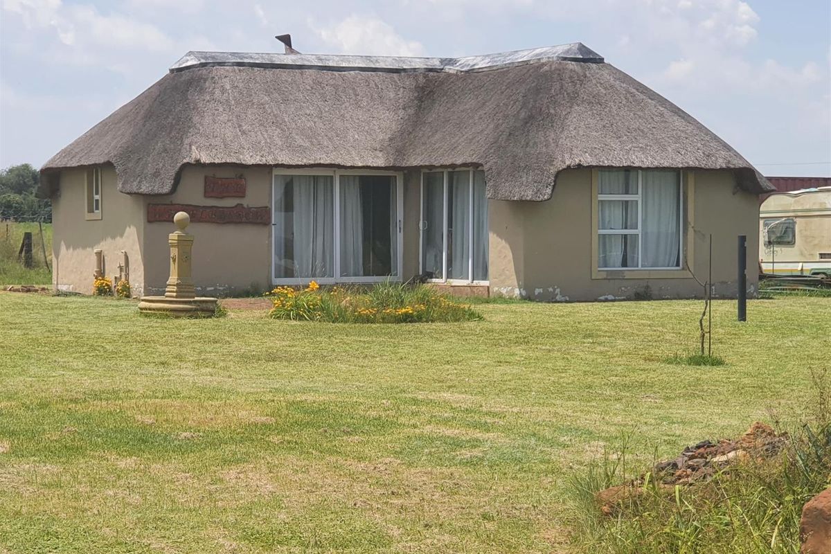 A single-story farmhouse featuring a traditional thatched roof, sliding glass doors, and a stone fountain set within a spacious garden.