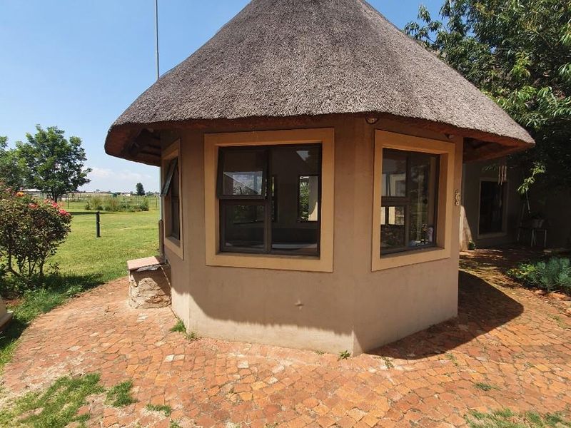 A charming thatched-roof gazebo structure featuring plastered walls and casement windows, set within a garden area with brick-paved surroundings.