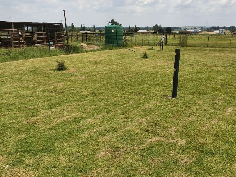 Expansive farm garden area featuring a green water storage tank, wire perimeter fencing, and a livestock enclosure in the background.
