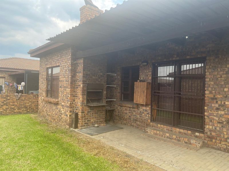 A covered outdoor patio area featuring a built-in brick braai, paved flooring, and security-fitted windows, overlooking a small garden space.