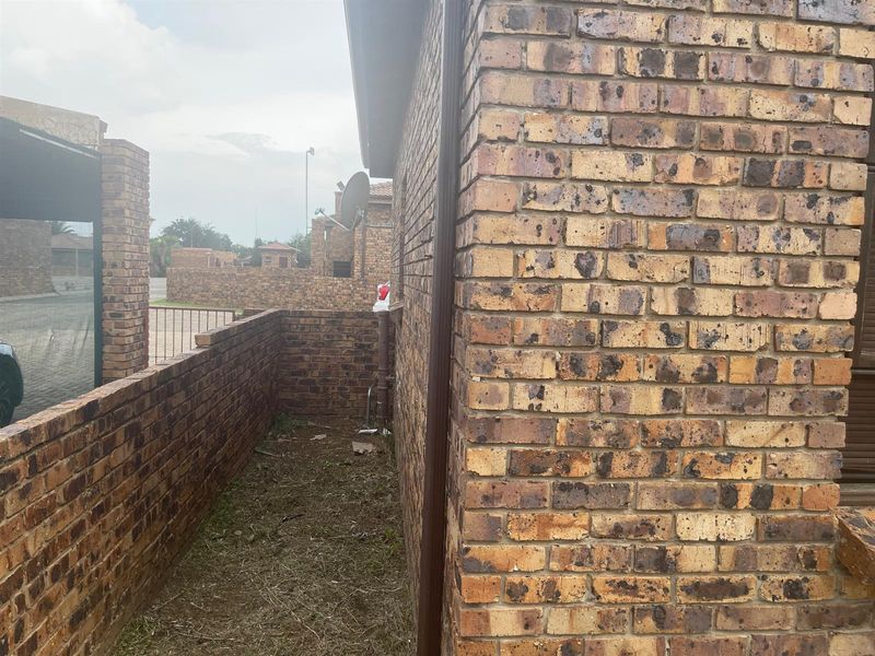 A side view of the property showing face brick exterior walls, a drainage downpipe, and a surrounding boundary wall with unmaintained ground.