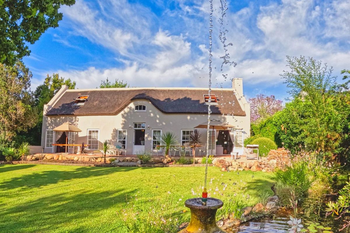 A charming thatched-roof house viewed from the rear garden, featuring a landscaped lawn, a decorative water fountain, and a paved patio area.