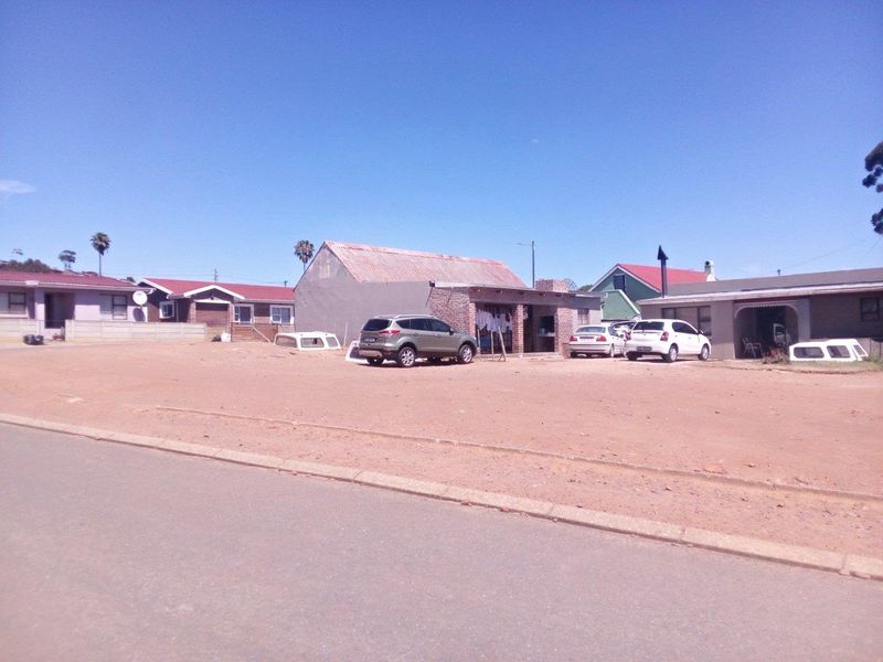 Street view of a residential property featuring an unpaved front yard area and existing dwelling structures with corrugated metal roofing.