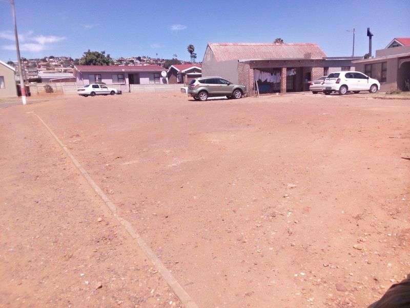 A vacant residential plot featuring an unpaved, open ground area with surrounding neighborhood residential structures visible in the background.