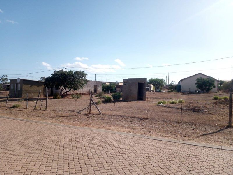 A view of a vacant residential plot featuring basic outbuildings, perimeter fencing, and established trees, accessible from a paved street.