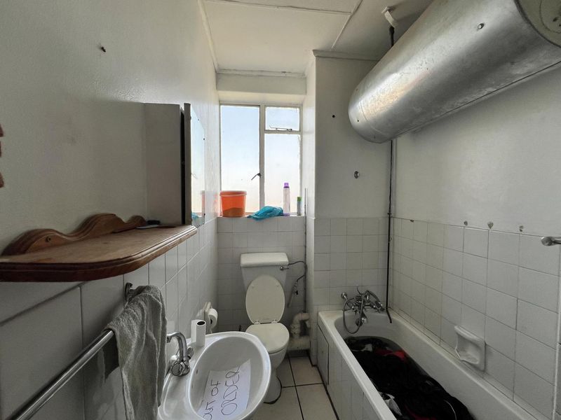 A dated bathroom featuring a standard bathtub, toilet, and wall-mounted basin, with an exposed water heater cylinder and original wall tiling.