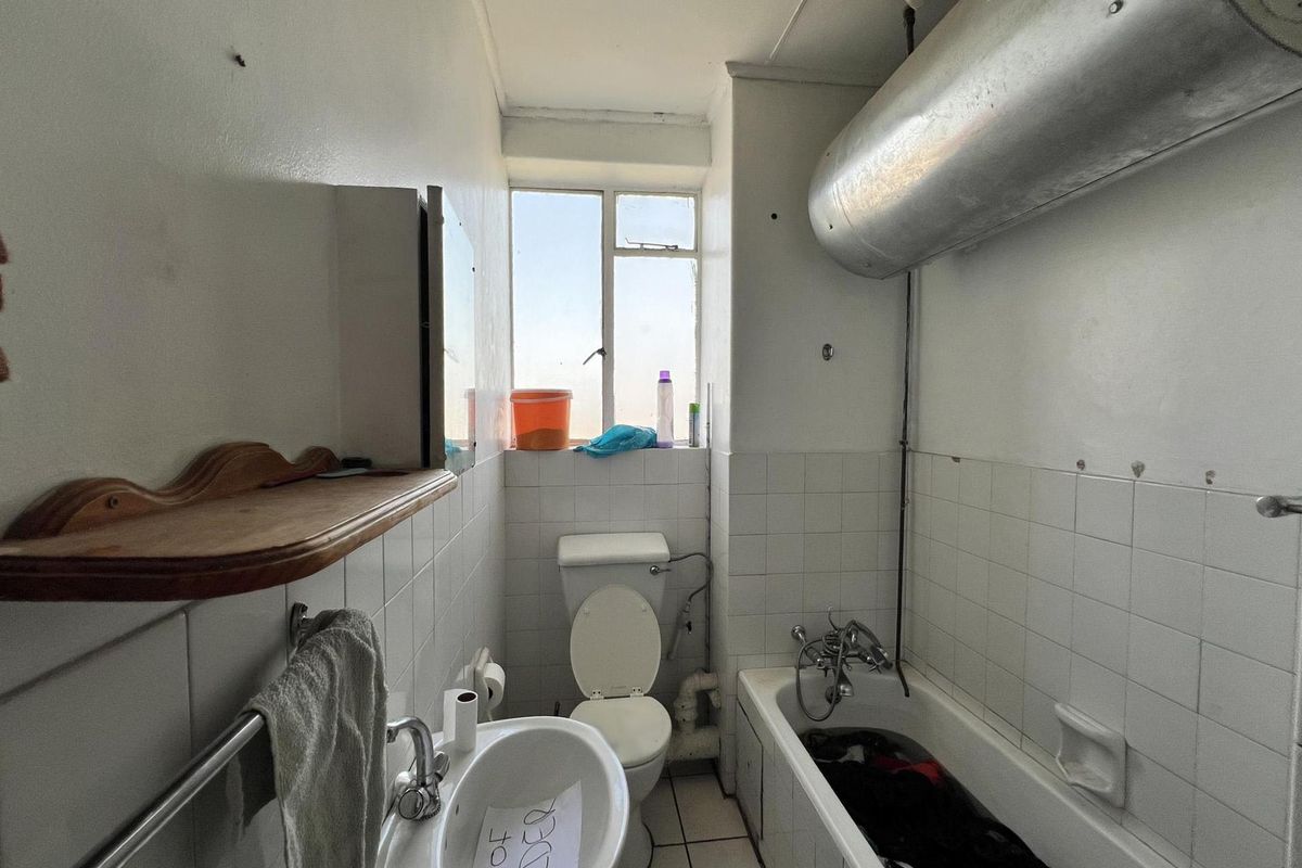 A dated bathroom featuring a standard bathtub, toilet, and wall-mounted basin, with an exposed water heater cylinder and original wall tiling.