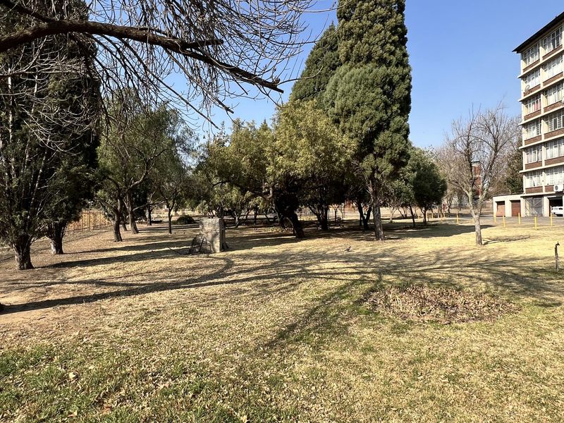 A view of the communal garden area featuring mature trees and dry lawn, adjacent to the townhouse complex building.