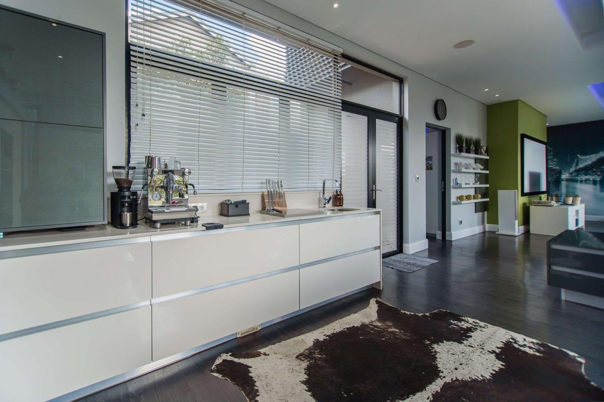 Modern kitchen area featuring sleek handleless cabinetry, integrated sink, and large windows with venetian blinds, flowing into an open-plan living space.