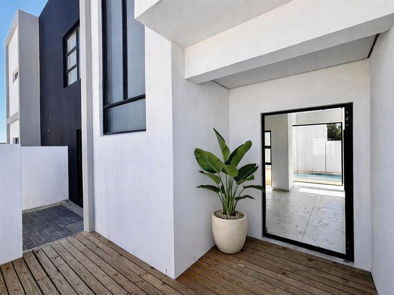 A modern covered patio area featuring timber decking, clean white architectural lines, and direct access to the pool area through a glass door.