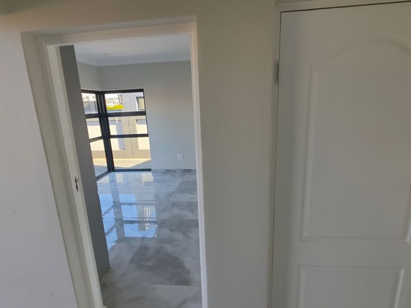 A bright, empty hallway featuring modern grey tiled flooring and clean white walls, leading into an adjacent room with large aluminium-framed windows.