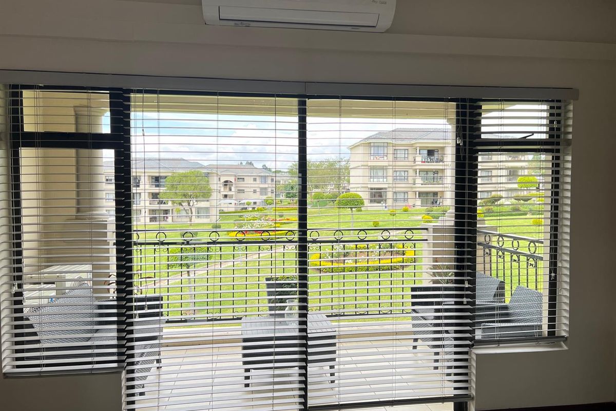 A view from an interior space looking out onto a balcony with a metal railing, overlooking a landscaped garden and apartment complex.