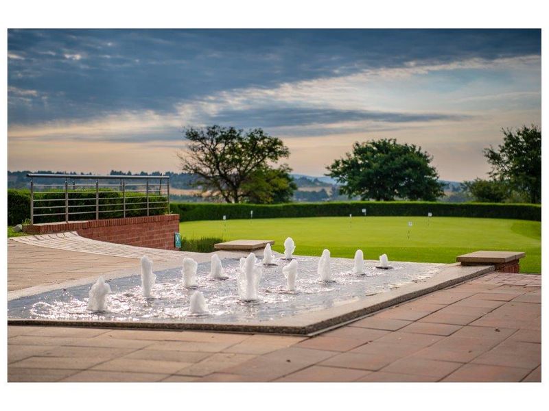 A scenic view of a well-maintained golf putting green featuring a decorative water fountain and paved patio area with brick retaining walls.