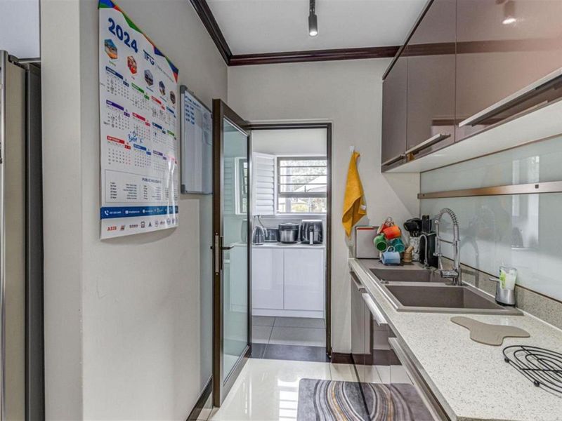 A functional scullery featuring quartz countertops, double basin sink, glossy cabinetry, and direct access to an adjacent utility area through a glass door.