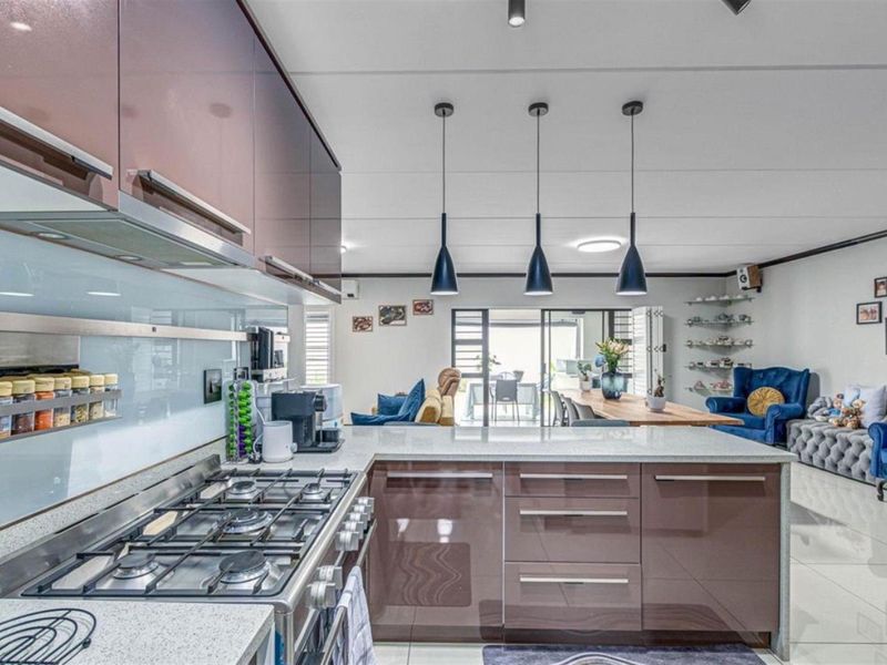 Modern open-plan kitchen and living area featuring high-gloss cabinetry, stone countertops, gas hob, and pendant lighting, flowing seamlessly to an outdoor patio space.