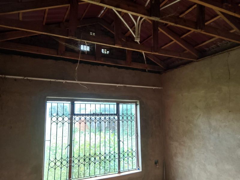An unfinished room featuring exposed timber roof trusses, plastered walls, and a security-fitted window, requiring significant renovation and ceiling installation.