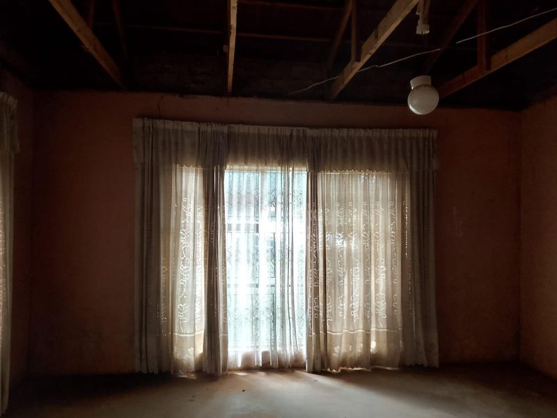 An empty living room featuring exposed timber ceiling beams and a window with security bars, requiring significant renovation and cosmetic updates.