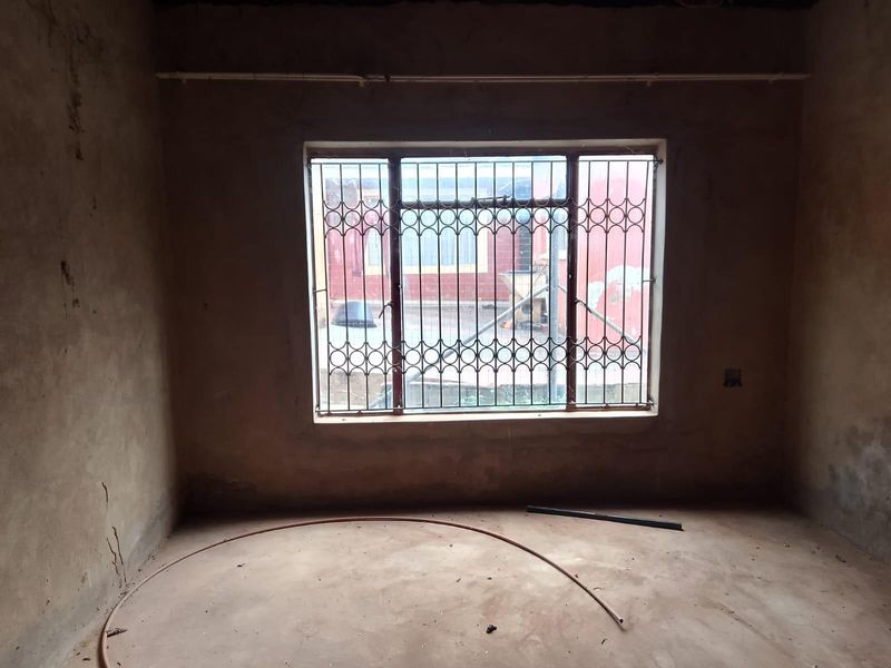 An unfinished bedroom space featuring bare plaster walls, concrete flooring, and a window fitted with security bars.