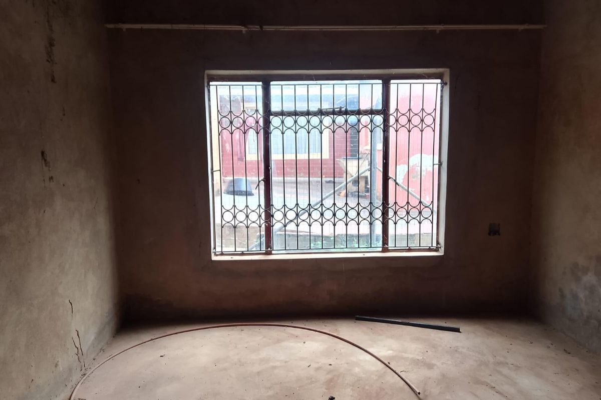 An unfinished bedroom space featuring bare plaster walls, concrete flooring, and a window fitted with security bars.