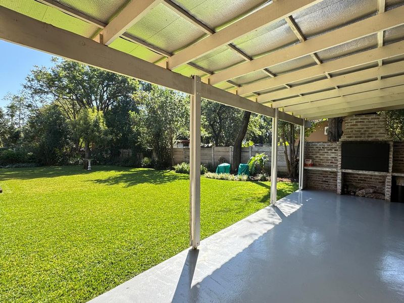 A spacious covered patio featuring a built-in brick braai, concrete flooring, and direct access to a well-maintained garden.
