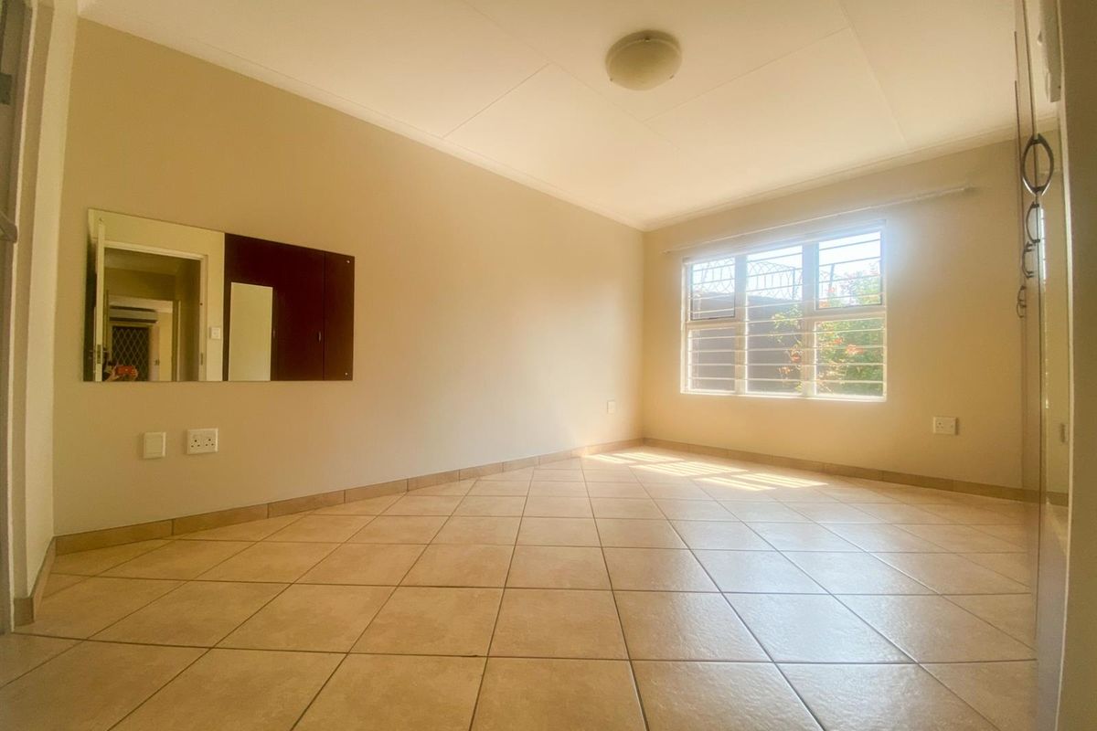 A bright, empty bedroom featuring neutral tiled flooring, a built-in wall mirror, and a window fitted with security bars.
