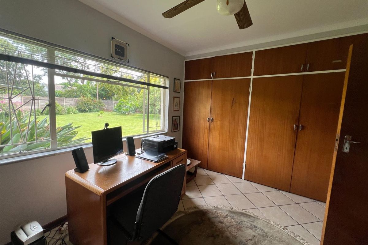 A functional bedroom featuring built-in wooden cupboards, tiled flooring, and a ceiling fan, with large windows overlooking the garden.