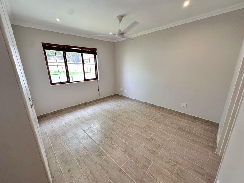 A bright, empty bedroom featuring modern wood-look tile flooring, a ceiling fan, recessed lighting, and a window with fitted wooden blinds.