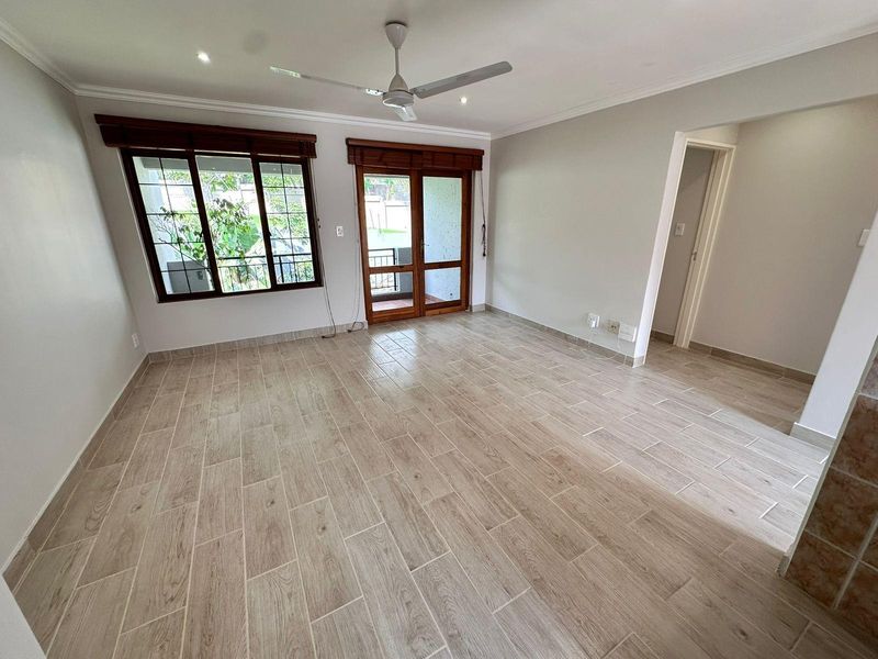 A bright, empty living room featuring modern wood-look tile flooring, a ceiling fan, and direct access to an outdoor balcony through timber-framed glass doors.