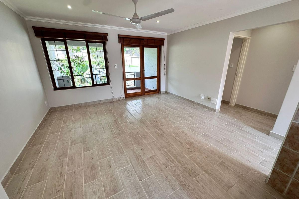 A bright, empty living room featuring modern wood-look tile flooring, a ceiling fan, and direct access to an outdoor balcony through timber-framed glass doors.