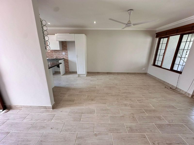 Spacious open-plan living area featuring modern wood-look tiled flooring, a ceiling fan, and an adjoining kitchen with granite countertops and white cabinetry.