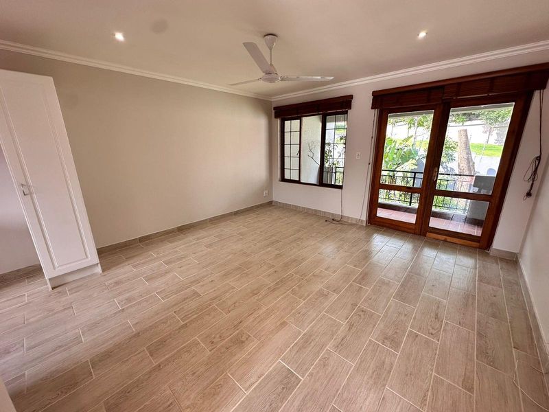 Spacious bedroom featuring wood-look tile flooring, built-in wardrobe, ceiling fan, and direct access to a private balcony through timber-framed glass doors.