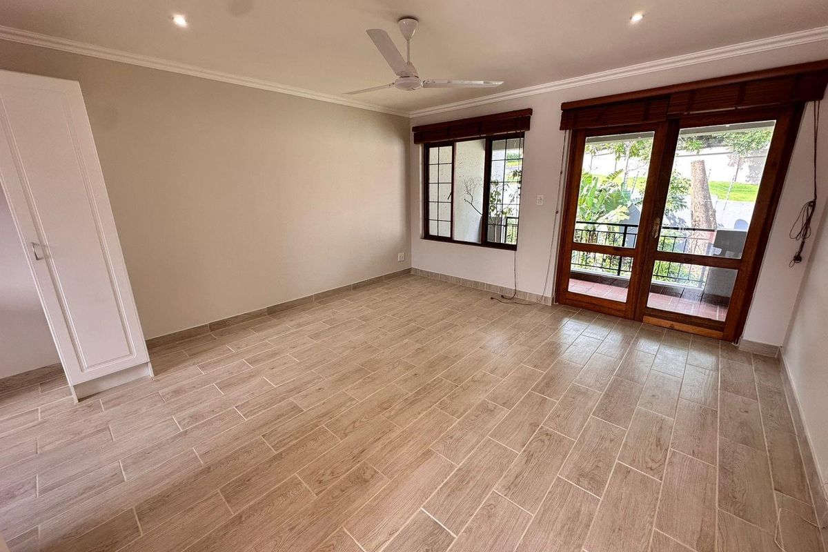 Spacious bedroom featuring wood-look tile flooring, built-in wardrobe, ceiling fan, and direct access to a private balcony through timber-framed glass doors.