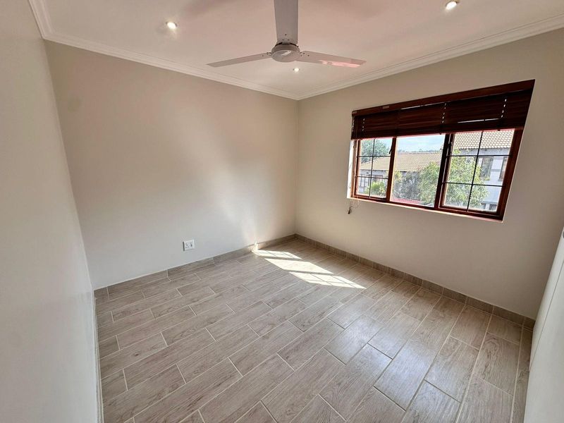 A bright, empty bedroom featuring modern wood-look tiled flooring, a ceiling fan, recessed lighting, and wooden blinds on the window.