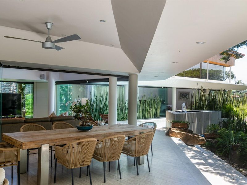 A spacious covered patio featuring a modern architectural design, recessed lighting, ceiling fan, and seamless transition to the landscaped garden and water feature.