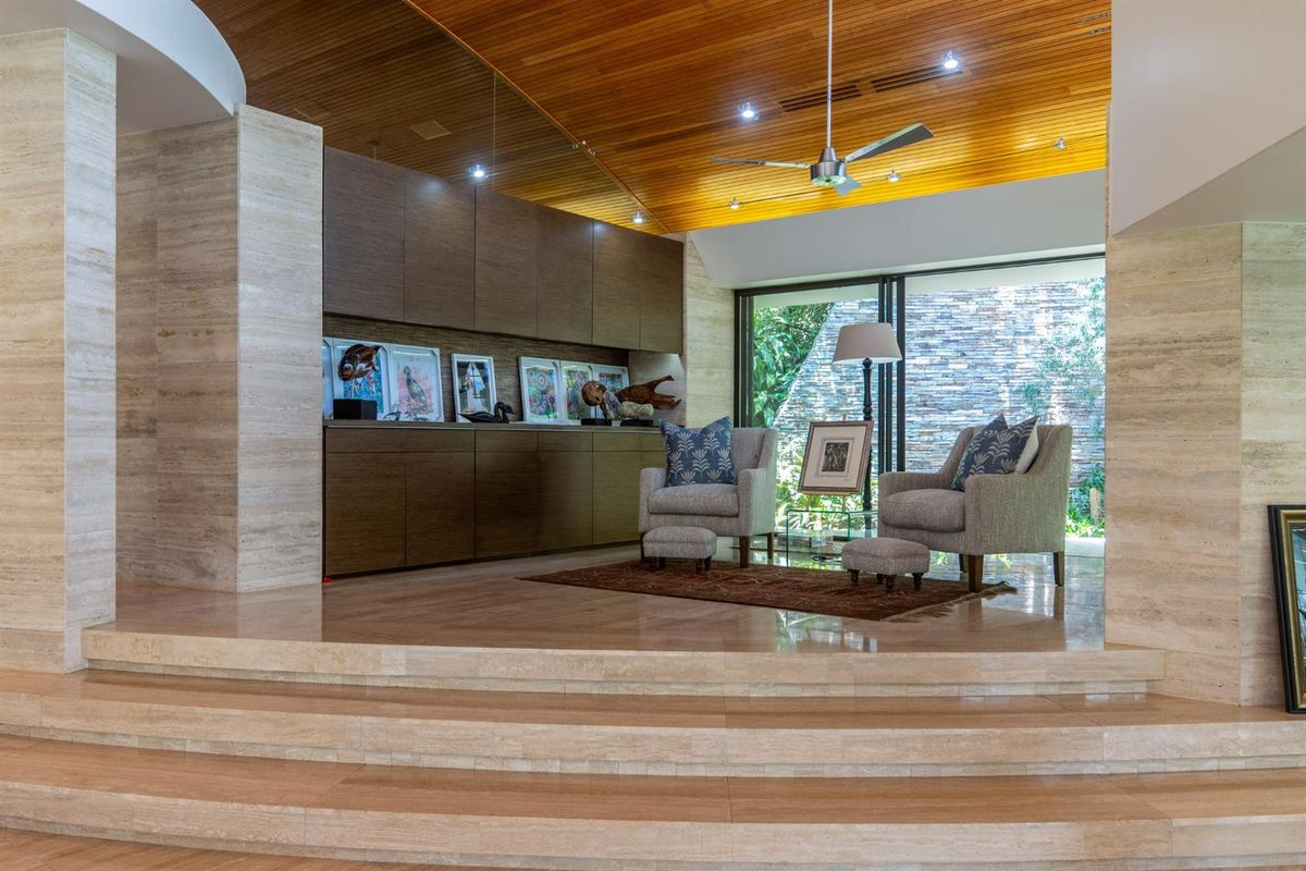 Spacious living area featuring high timber-paneled ceilings, travertine stone flooring, built-in cabinetry, and large sliding glass doors opening to a private garden view.