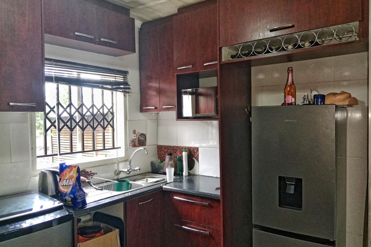 A functional kitchen featuring dark wood-toned cabinetry, laminate countertops, a stainless steel sink, and a tiled backsplash with a window security gate.