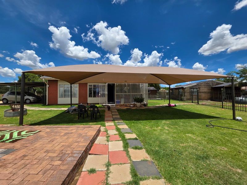 Rear exterior view of the property featuring a large shade sail, brick paving, and a well-maintained lawn area.