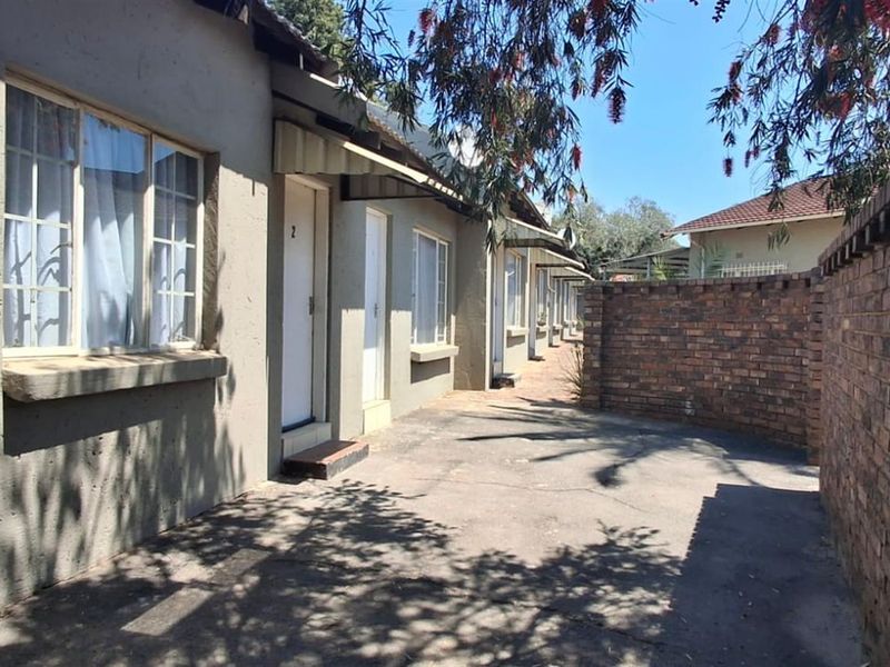 Exterior view of a residential unit row featuring a paved walkway, brick boundary wall, and multiple entrance doors with window awnings.