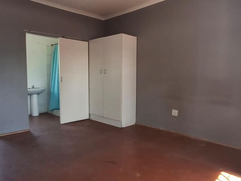 An open-plan living area featuring a basic kitchen sink, built-in wardrobe, and an adjoining bathroom with a pedestal basin and shower.