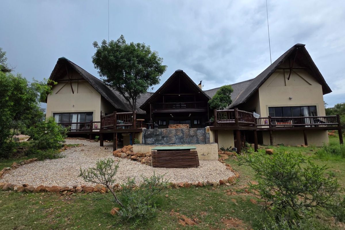 A spacious rear exterior view of a large house featuring a thatched roof, expansive wooden decking, and a central stone-clad water feature.